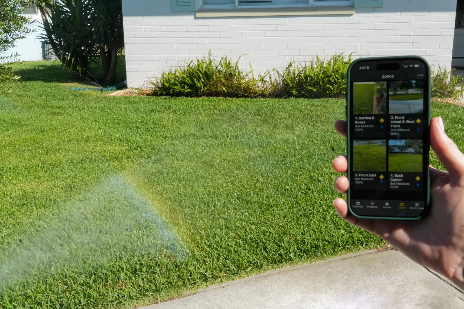 Homeowner adjusting irrigation zones from a smartphone app while a WaterCare-installed zone runs on the lawn behind